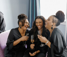 Three women in black sit on a purple couch, laughing with champagne glasses—an elegant Gulf South wedding inspiration moment.