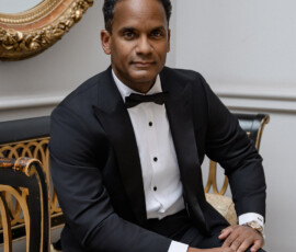 Man in black tuxedo sits on ornate chair before a gold-framed mirror, exuding New Orleans wedding elegance.