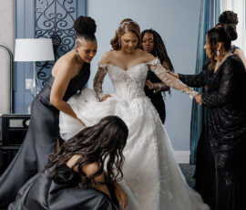 Bride in white gown with bridesmaids in black dresses adjusting her dress and veil in an elegant, softly lit New Orleans room.