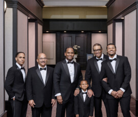 Six men and boys in black tuxedos pose, smiling in an elegant hallway at a Louisiana wedding with ornate ceilings and soft lighting.