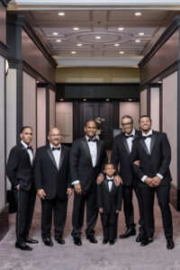 Groom poses with father, grandfather, groomsmen, and young ring bearer in matching black tuxedos in elegant hotel hallway