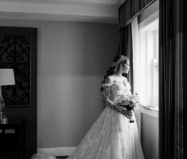 Bride in lace gown stands by window with bouquet in softly lit New Orleans room, a scene of timeless wedding inspiration.
