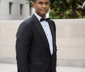 Man in black tuxedo and bow tie stands outside near greenery and modern building at a Louisiana wedding.