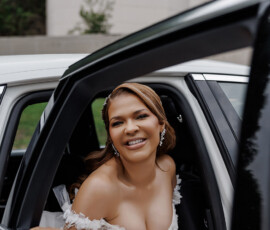 Smiling bride in off-shoulder white dress sits in car with door open, joyfully looking out before her Louisiana wedding.
