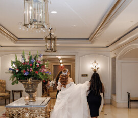 Bride in white gown walks down elegant hallway with helper, surrounded by florals, chandeliers, and classic wedding decor.