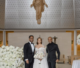 Bride in white gown with two men in black suits poses before Jesus statue amid white flowers in church wedding scene.