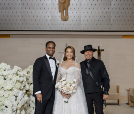 Bride in white gown smiles between two men in a church with white flowers and a Jesus statue at a joyful Louisiana wedding.