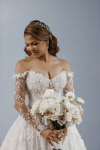 Bridal portrait of bride in floral lace off-the-shoulder wedding gown holding white rose bouquet against soft blue background