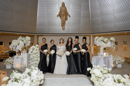 Bride and bridesmaids in black satin dresses pose inside church ceremony space surrounded by white florals and candlelit aisle décor