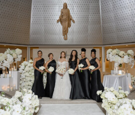 Bride in white gown with five bridesmaids in black, holding white bouquets, in a classic New Orleans church with candles and flowers.