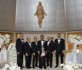 Groom, groomsmen, and a boy in tuxedos pose in a church with white flowers and candles under a statue of Jesus.