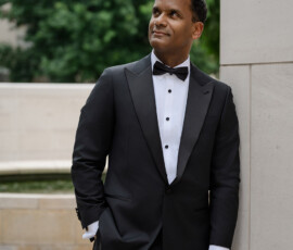 Man in black tuxedo and bow tie leans on stone wall outdoors, smiling with trees behind—ideal Gulf South wedding inspiration.