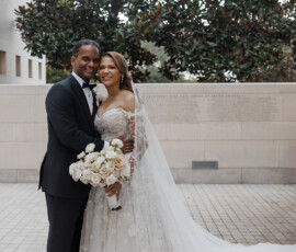 Bride in lace gown with white roses and groom in tuxedo smile by tree and stone wall at a Louisiana wedding.