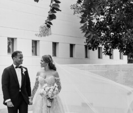 Bride and groom holding hands outdoors at their Louisiana wedding, smiling, with trees and a modern building in the background.