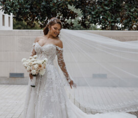 Bride in lace wedding gown with train and veil, holding pastel rose bouquet outdoors by tiled surface and greenery.