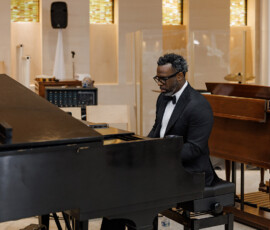 A man in a tuxedo plays a grand piano in a bright room with beige walls and glass panels, perfect for New Orleans wedding inspiration.