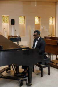 Pianist in black tuxedo playing grand piano during wedding ceremony inside modern church with warm candlelight and white floral décor