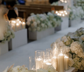 White candles in glass holders and white flowers line the aisle at an elegant Louisiana indoor wedding ceremony.