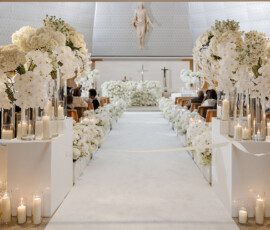 Wedding aisle with white flowers and candles in a church, leading to a flower-adorned altar and statue above.