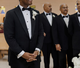 Smiling man in black tuxedo stands at church front with three groomsmen and white flowers, classic Louisiana wedding.