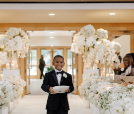 Smiling young boy in black tuxedo carries ring pillow down flower-lined aisle at New Orleans wedding as guests look on.