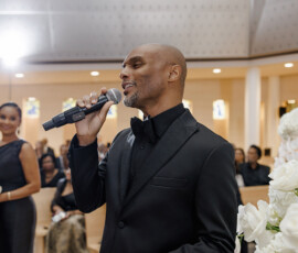 Man in black tuxedo sings at indoor wedding reception, guests seated behind him and white flowers in front.