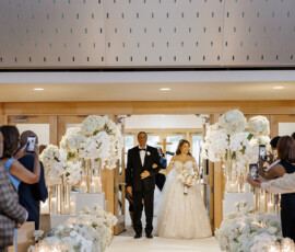 Bride in white gown walks down aisle with man in tux at elegant Louisiana wedding, guests capturing the flower-filled scene.