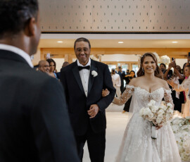 Bride in off-shoulder gown walks arm-in-arm with tuxedoed man down aisle at elegant New Orleans wedding, guests watching.