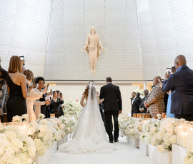 Bride in white dress walks down flower-filled church aisle in New Orleans wedding under statue of Jesus, guests taking photos.