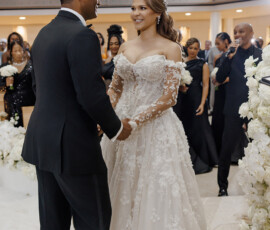 Bride in lace gown and groom in tuxedo smile, holding hands at their New Orleans wedding with guests and white flowers.