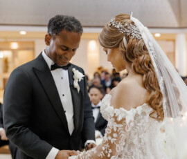 Bride in lace gown and groom in black tuxedo hold hands at a Louisiana wedding ceremony, guests seated behind them.