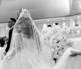 Bride in lace gown and veil walks aisle, holding train; groom nearby, white flowers decorate black-and-white Louisiana wedding.