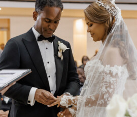 Groom in black tux places ring on bride in lace gown at indoor New Orleans wedding, inspiring couples’ celebrations.