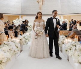 Bride and groom walk hand in hand, smiling down the aisle of a flower-filled Louisiana church as guests celebrate and take photos.