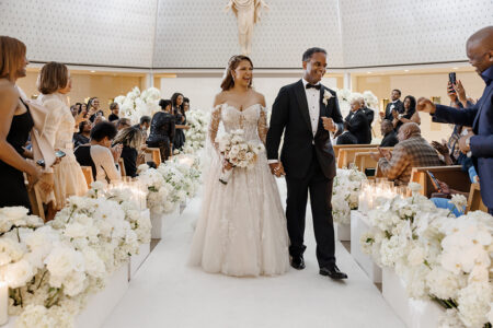 Newlyweds walk down a flower-lined aisle hand in hand after the ceremony as guests applaud from their seats.