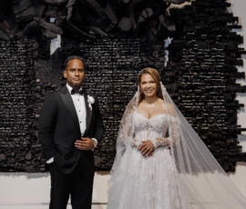 Bride in white gown and veil stands with groom in tuxedo indoors before Louisiana wedding, against dark textured wall art.