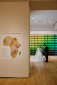 Bride and groom stand inside a contemporary art gallery framed by a glowing green installation and museum exhibits.