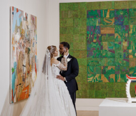 Bride and groom embrace in a New Orleans art gallery, surrounded by abstract paintings; she wears white gown, he a dark suit.