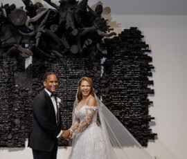 Bride in lace gown and groom in black tux smile, holding hands before sculptural wall at an elegant Louisiana wedding.