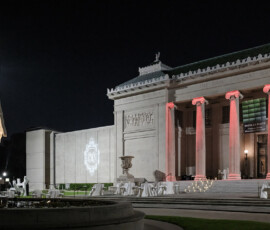 Elegant night view of a grand museum with red-lit columns, outdoor staircase, sculpture, and projected logo—ideal for weddings.