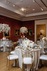 Reception dining room with cream linens, tall floral centerpieces, candlelight, and gold geometric chairs surrounded by artwork.