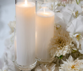Lit glass candles with white wax, surrounded by white and pale pink dahlias and orchids for a serene wedding display.