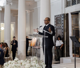 Man in suit speaks at clear podium in elegant room with columns and flowers at Louisiana wedding; guests listen in background.