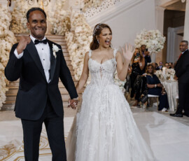 Smiling bride and groom walk hand in hand at their New Orleans wedding reception, waving amid guests and floral decor.