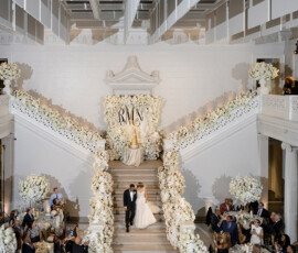 Bride and groom descend flower-lined stairs at an elegant indoor Louisiana wedding with lush white decor and a monogrammed floor.