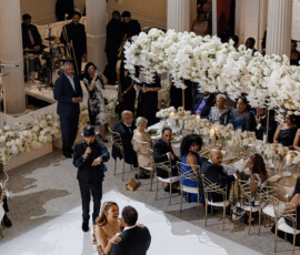 Bride and groom share first dance in elegant hall with white columns, floral decor, and guests at round tables.