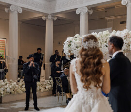 Bride and groom face a band at an elegant indoor wedding with white flowers, tall columns, and guests viewing from above.