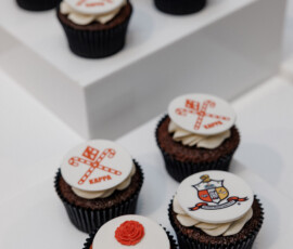 Chocolate cupcakes with white icing and red KAPPA designs, a rose with 1913, and shield emblems on elegant white stands.