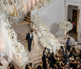 Bride and groom descend a flower-lined staircase as guests take photos at an elegant, gold-accented New Orleans wedding.