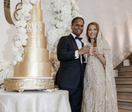 Couple in formal wear smiles by tall gold wedding cake, holding champagne, with floral arch and RN initials at Gulf South reception.